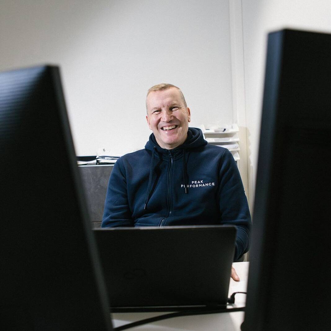 Hymyilevä henkilö istuu toimistossa kahden tietokonenäytön välissä. Hänellä on tummansininen huppari, jossa lukee 