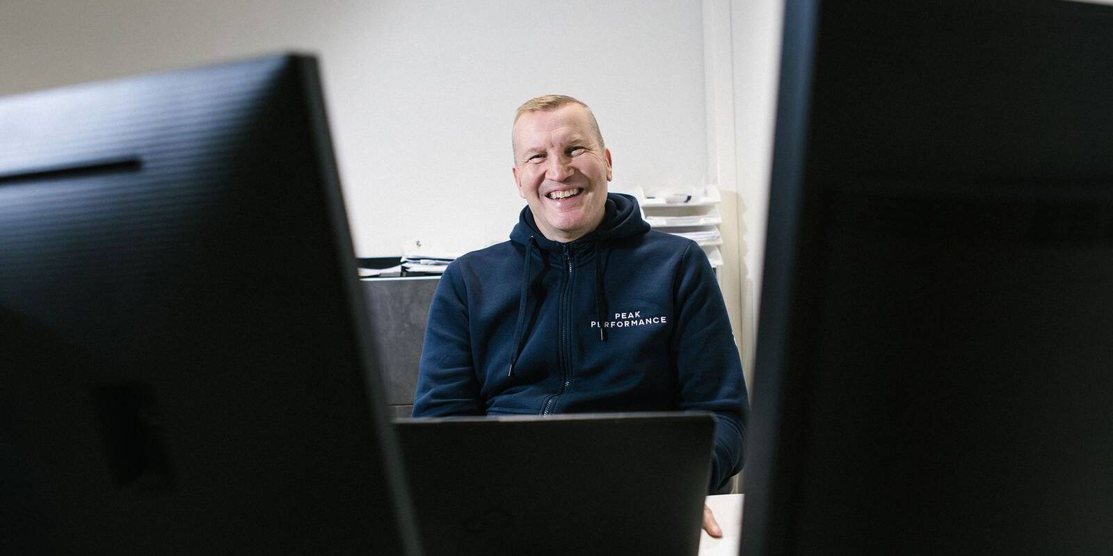Hymyilevä henkilö istuu toimistossa kahden tietokonenäytön välissä. Hänellä on tummansininen huppari, jossa lukee "Peak Performance". Taustalla näkyy valkoinen seinä ja pino papereita. Kuva välittää rennon ja hyväntuulisen tunnelman työpäivän keskeltä.