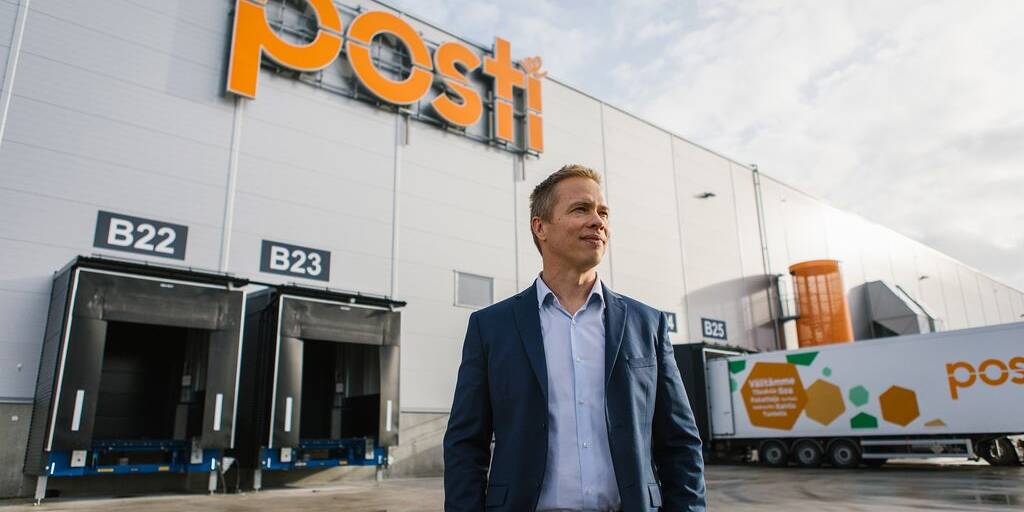 Pukumies seisoo Postin logistiikkakeskuksen edessä; taustalla näkyvät lastauslaiturit ja Postin brändätty rekka.