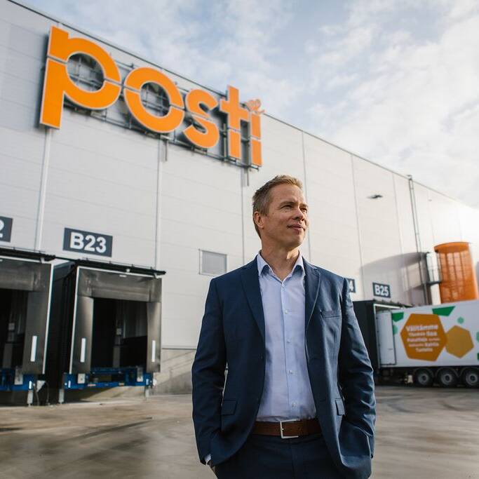 Pukumies seisoo Postin logistiikkakeskuksen edessä; taustalla näkyvät lastauslaiturit ja Postin brändätty rekka.