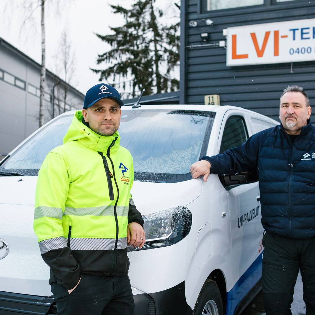 Kaksi LVI-alan työntekijää poseeraa talvisessa säässä yrityksen pakettiautojen edessä. Toisella on päällään kirkkaan keltainen huomiotakki ja toisella tummansininen työasu. Taustalla näkyy LVI-tekniikkaa tarjoavan yrityksen rakennus ja kyltti.