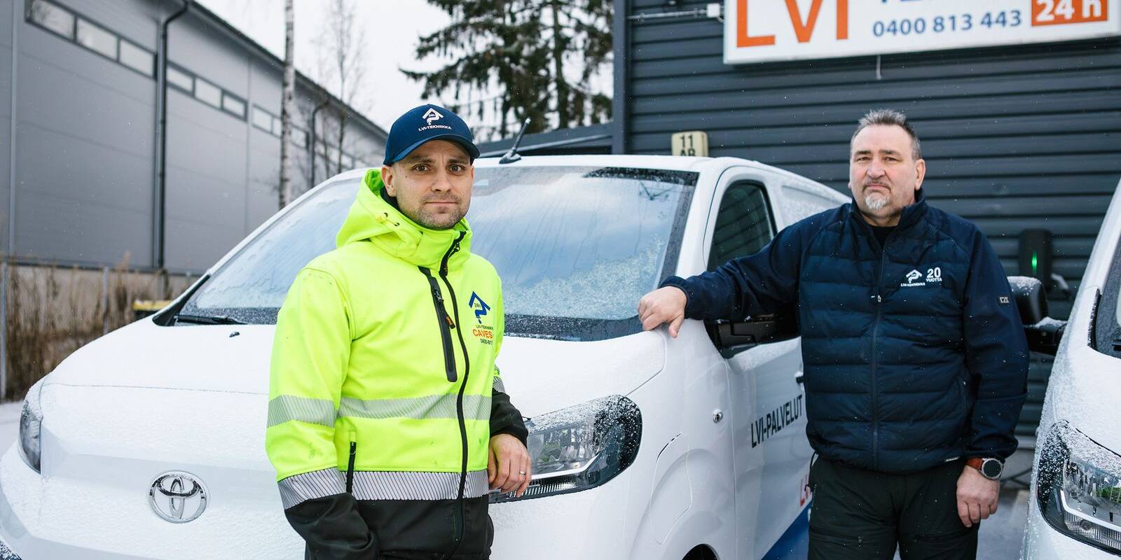 Kaksi LVI-alan työntekijää poseeraa talvisessa säässä yrityksen pakettiautojen edessä. Toisella on päällään kirkkaan keltainen huomiotakki ja toisella tummansininen työasu. Taustalla näkyy LVI-tekniikkaa tarjoavan yrityksen rakennus ja kyltti.