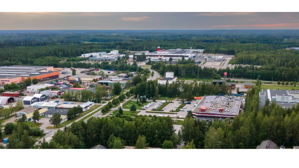 Ilmakuva teollisuusalueesta metsän keskellä. Kuvassa näkyy useita suuria hallirakennuksia, pysäköintialueita ja teitä. Taustalla avautuu laaja metsäinen maisema ja horisontissa iltataivas.