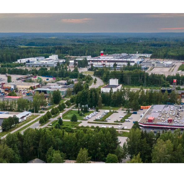 Ilmakuva teollisuusalueesta metsän keskellä. Kuvassa näkyy useita suuria hallirakennuksia, pysäköintialueita ja teitä. Taustalla avautuu laaja metsäinen maisema ja horisontissa iltataivas.