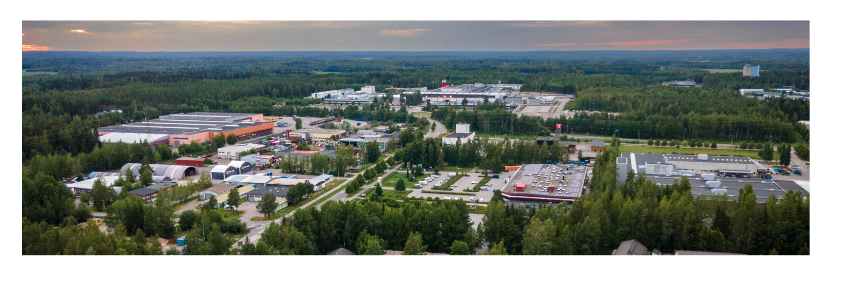 Ilmakuva teollisuusalueesta metsän keskellä. Kuvassa näkyy useita suuria hallirakennuksia, pysäköintialueita ja teitä. Taustalla avautuu laaja metsäinen maisema ja horisontissa iltataivas.