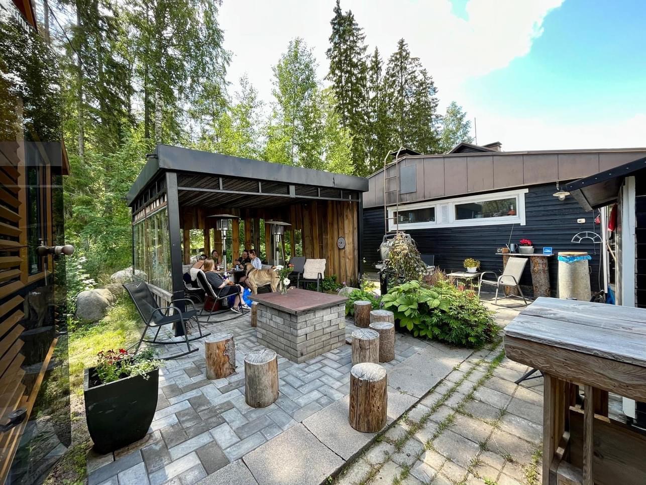 Suojaisa grillikatos pihapiirissä metsän keskellä, ihmiset istuvat pöydän ääressä ja keskellä on tiilinen tulipaikka.