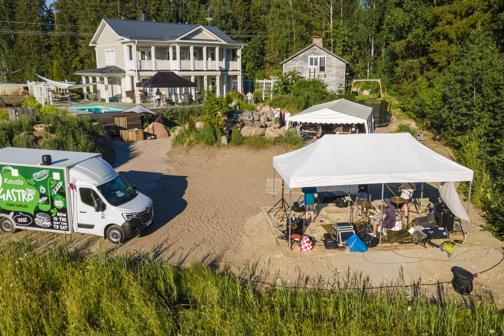 Ilmakuva suuresta vaaleasta huvilasta pihapiireineen, pihalla juhlateltta ja lava, vieressä ruokarekka ja metsää ympärillä.