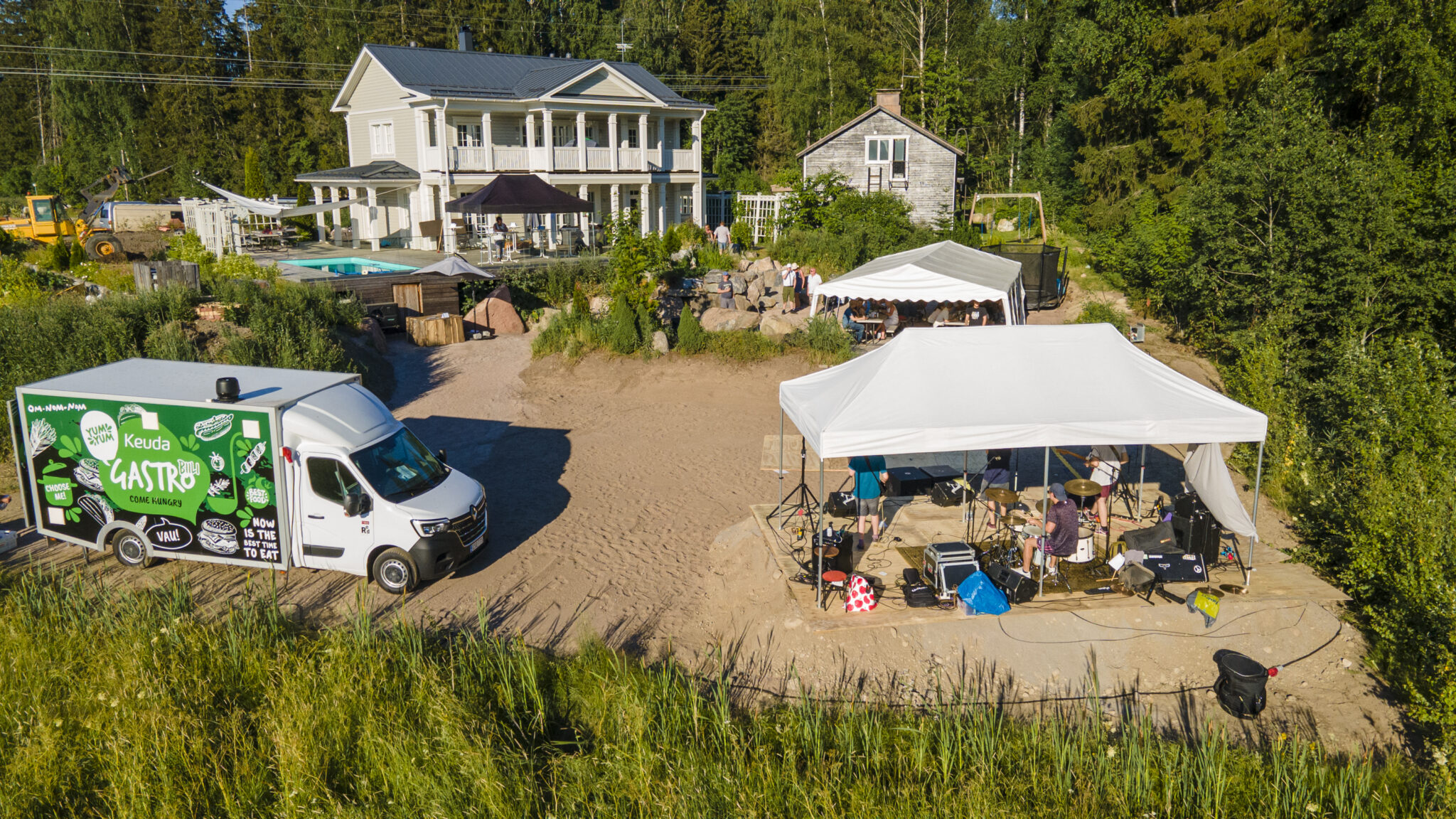Ilmakuva suuresta vaaleasta huvilasta pihapiireineen, pihalla juhlateltta ja lava, vieressä ruokarekka ja metsää ympärillä.