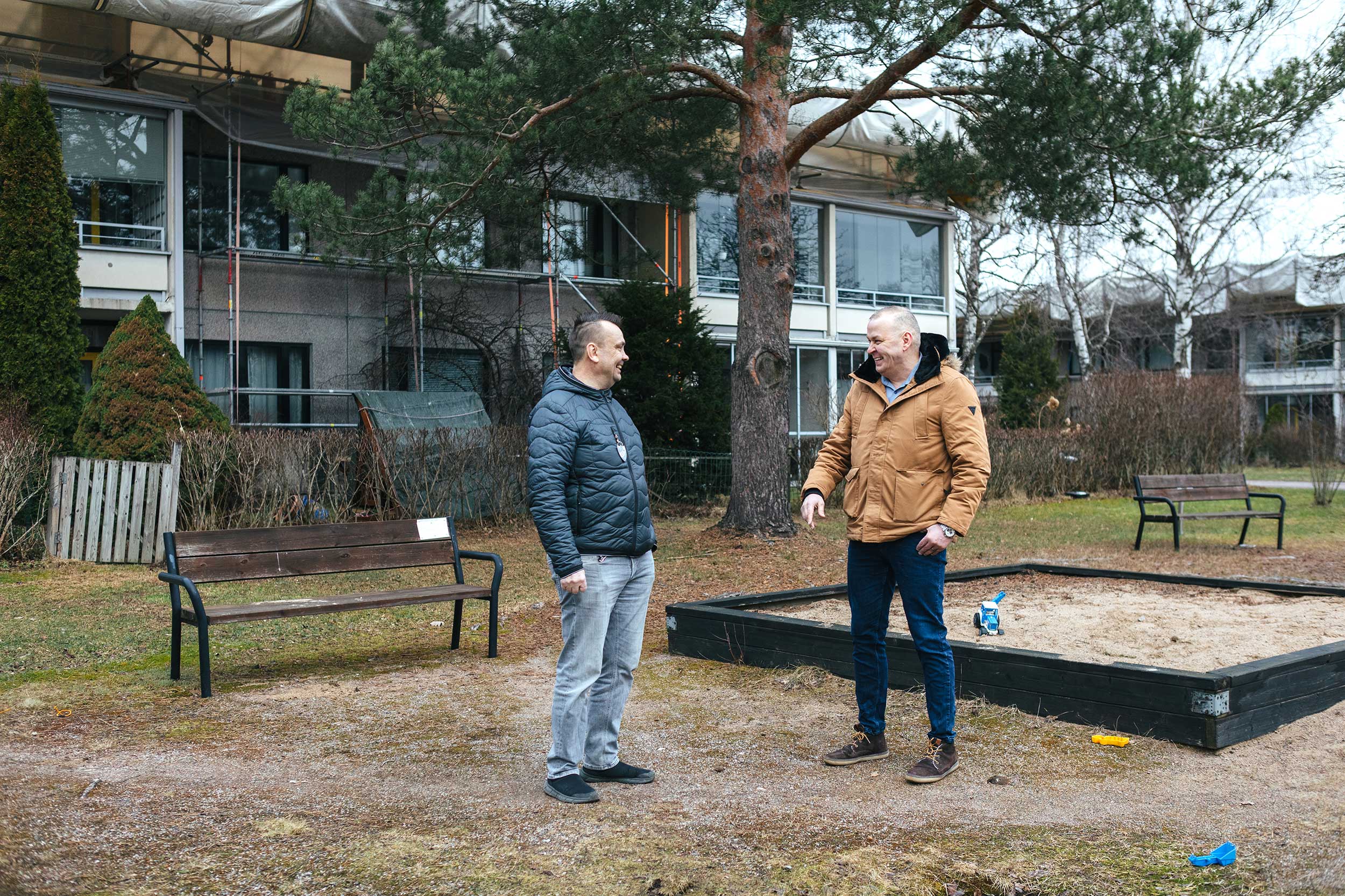 Kaksi miestä keskustelee ulkona pihalla kerrostalon edustalla. He seisovat hiekkalaatikon ja penkkien lähellä, taustalla näkyy puita ja asuinrakennuksia.