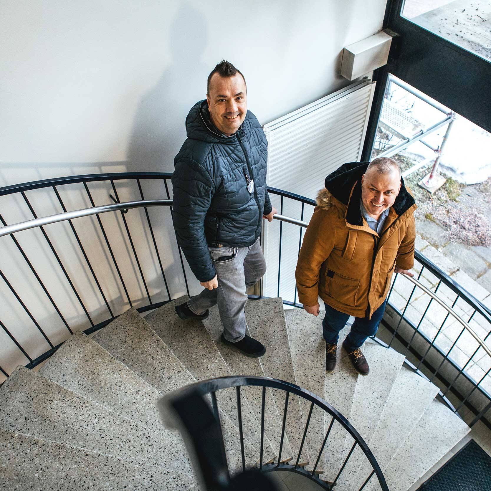 Kaksi miestä kävelee spiraalimaisia portaita alas sisätiloissa. He katsovat ylöspäin kameraan ja hymyilevät. Tilassa on vaalea seinä, kaide ja suuri ikkuna, josta näkyy ulkona maata ja hieman lunta.