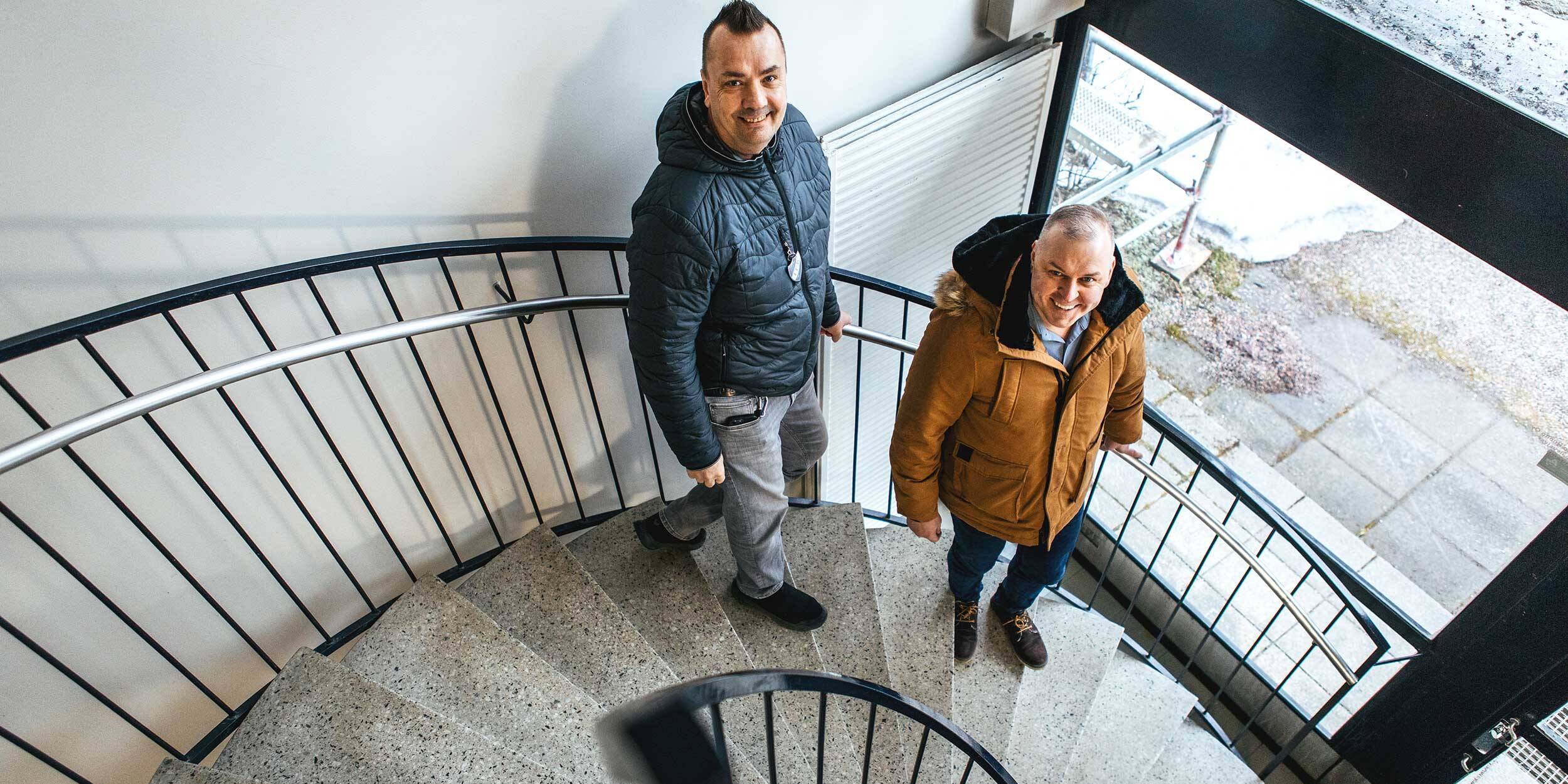 Kaksi miestä kävelee spiraalimaisia portaita alas sisätiloissa. He katsovat ylöspäin kameraan ja hymyilevät. Tilassa on vaalea seinä, kaide ja suuri ikkuna, josta näkyy ulkona maata ja hieman lunta.