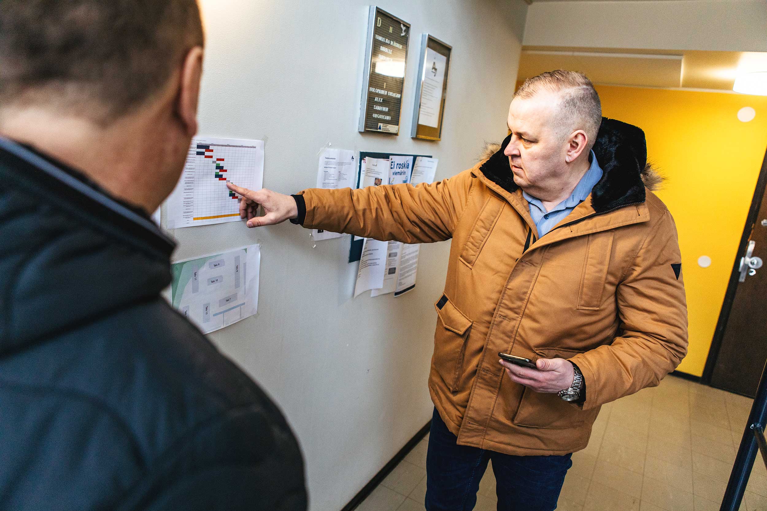 Kaksi miestä seisoo sisätilassa ilmoitustaulun äärellä. Toinen osoittaa seinällä olevaa kaaviota ja pitää toisessa kädessään puhelinta, kun taas toinen katsoo vierestä. Taustalla näkyy erilaisia ilmoituksia ja käytävä.