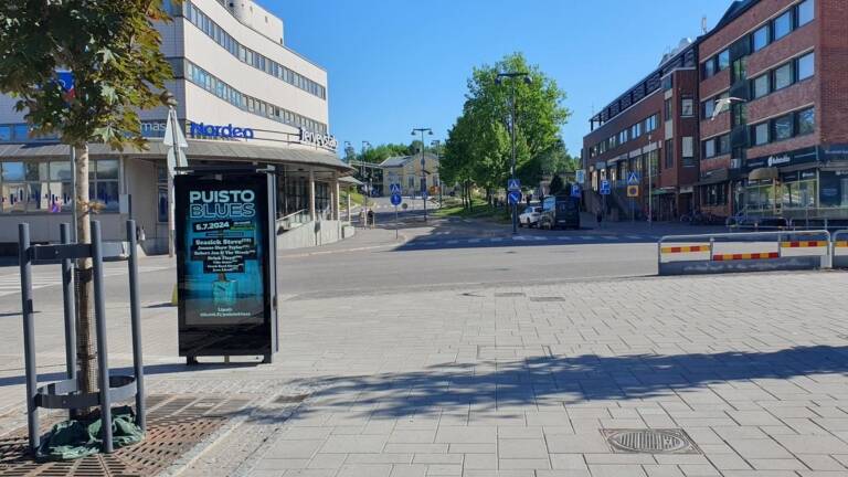 Aurinkoinen kaupunkinäkymä leveältä kävely- ja katualueelta. Vasemmalla vaalea liikerakennus, jonka edessä mainostaulu “Puisto Blues” -tapahtumasta. Oikealla punatiilinen rakennusrivi, katutasossa liiketiloja. Keskellä risteys, muutama pysäköity auto ja liikennemerkkejä, taustalla puita ja loiva mäki.