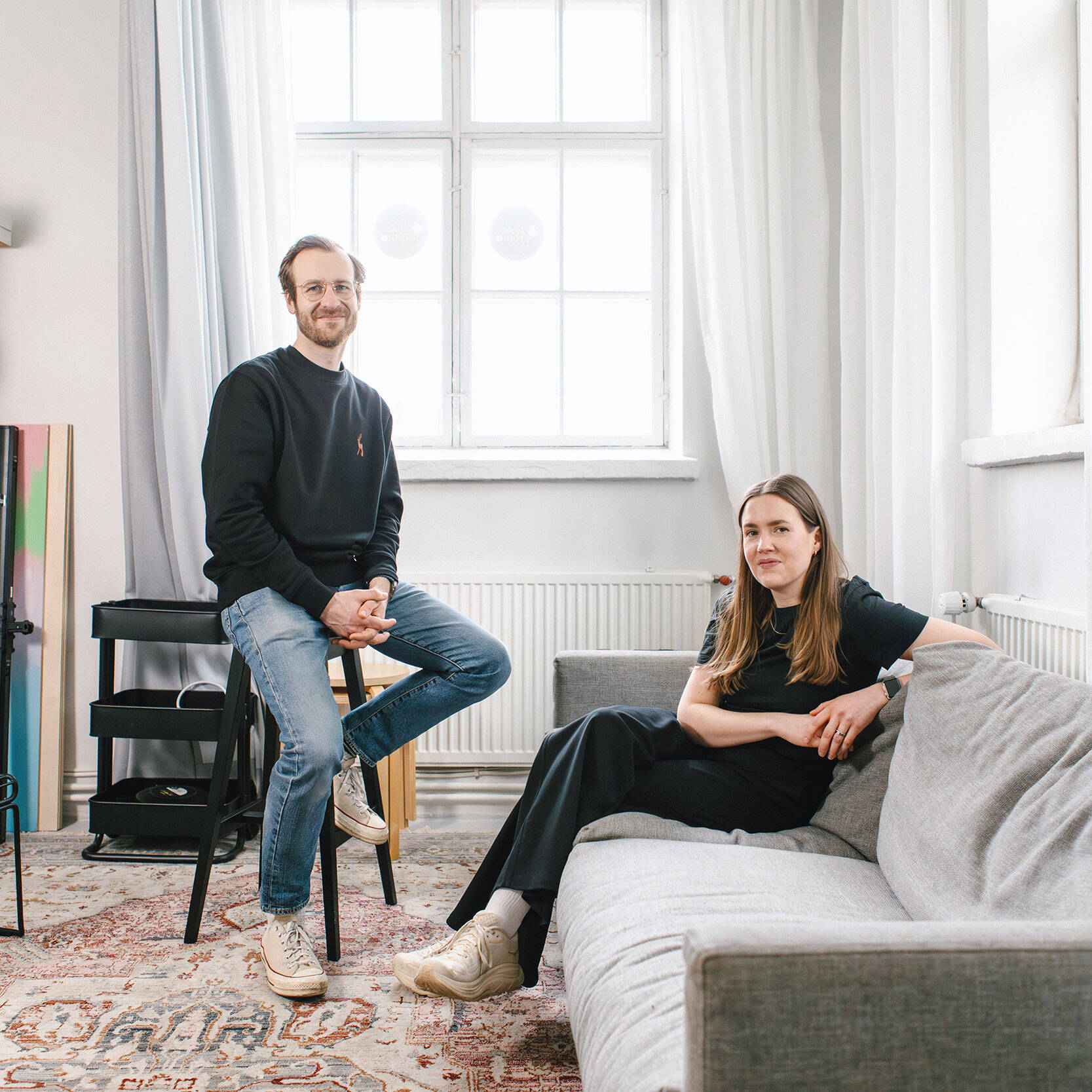 Kaksi henkilöä oleskelee valoisassa huoneessa. Toinen istuu korkealla jakkaralla ja toinen sohvalla. Taustalla on iso ikkuna, hylly ja kuvausvälineitä. Tunnelma on rento ja he katsovat kameraan.