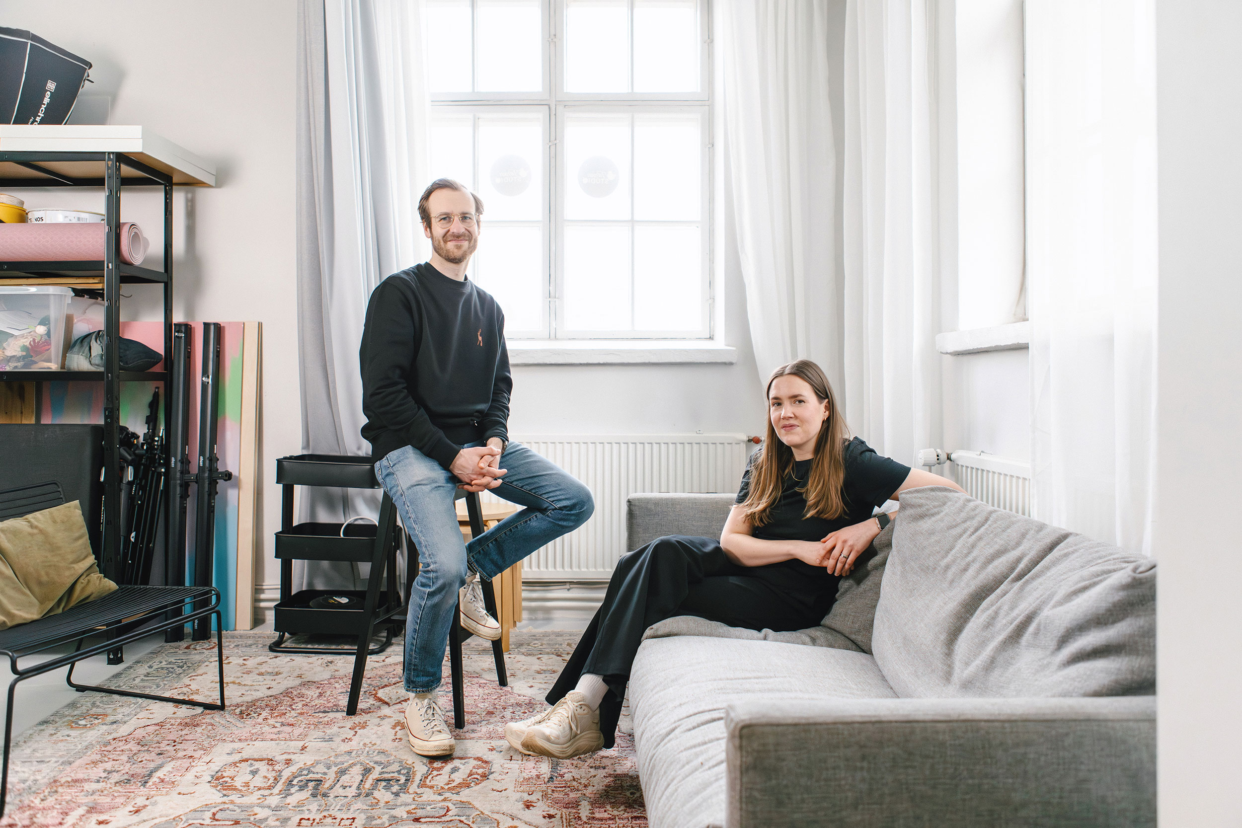 Kaksi henkilöä oleskelee valoisassa huoneessa. Toinen istuu korkealla jakkaralla ja toinen sohvalla. Taustalla on iso ikkuna, hylly ja kuvausvälineitä. Tunnelma on rento ja he katsovat kameraan.