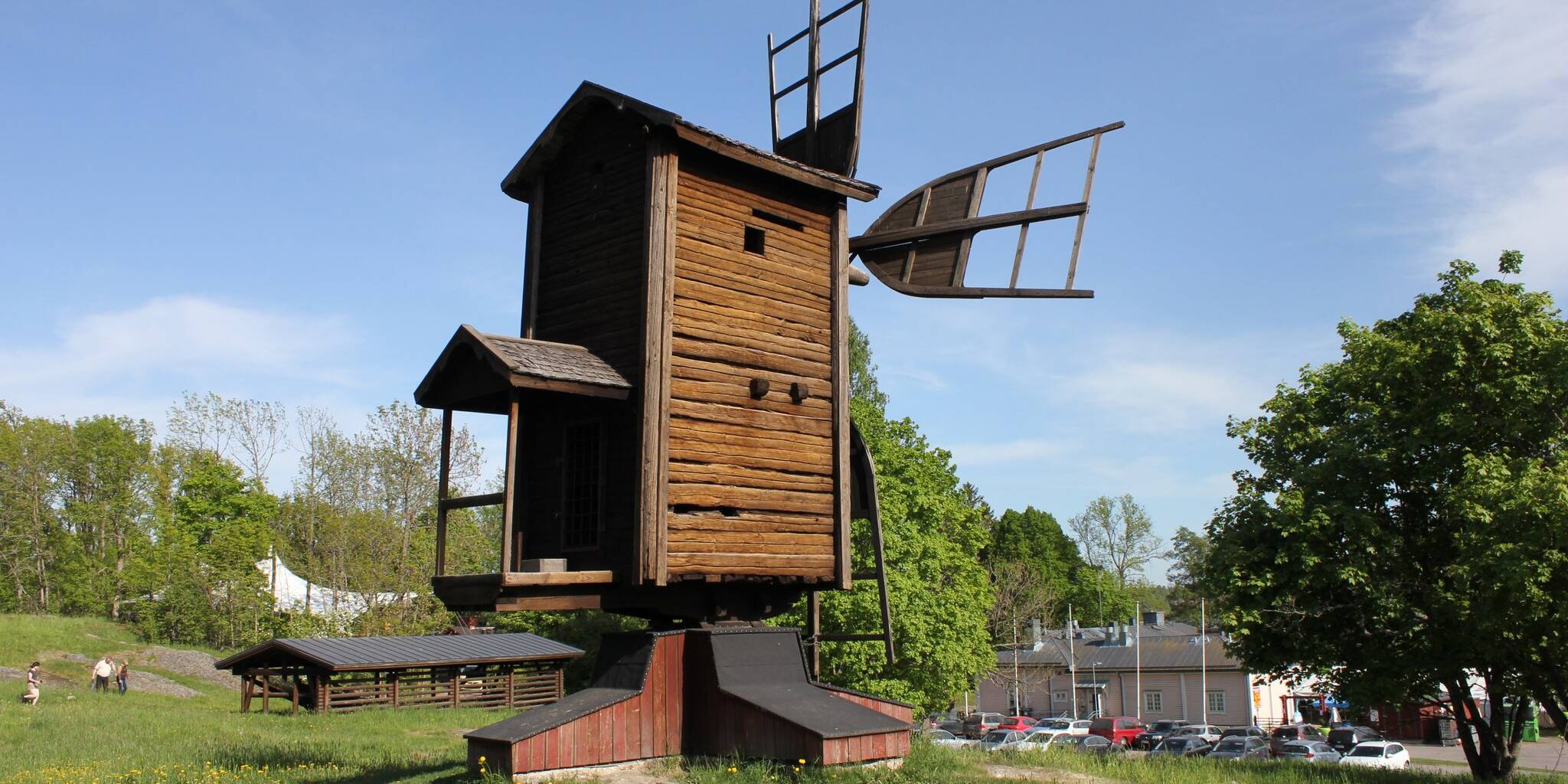 Puinen tuulimylly aurinkoisella nurmialueella. Myllyn siivet ovat osittain näkyvissä, ja taustalla näkyy puita, rakennuksia sekä sininen taivas ohuine pilvineen.