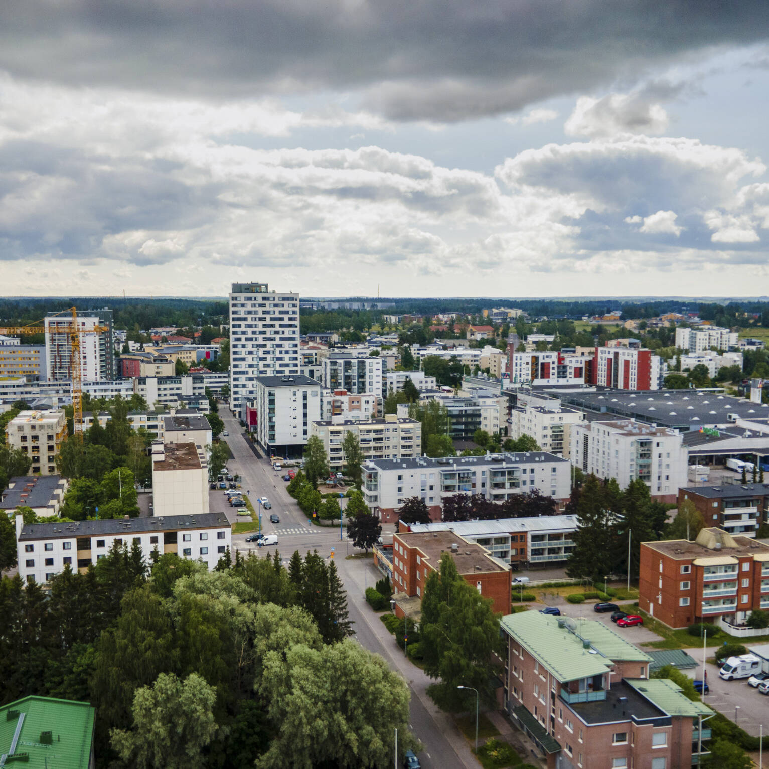 Ilmakuva Järvenpään keskustasta pilvisenä kesäpäivänä. Kuvassa näkyy tiivistä kaupunkirakennetta, kerrostaloja, liikekeskuksia ja vihreitä puistoalueita. Keskellä kohoaa useita korkeita asuinrakennuksia, ja etualalla näkyy matalampia punatiilisiä kerrostaloja puiden ympäröiminä. Taivaalla on suuria tummia pilviä, joiden välistä siivilöityy auringonvaloa kaupungin ylle.