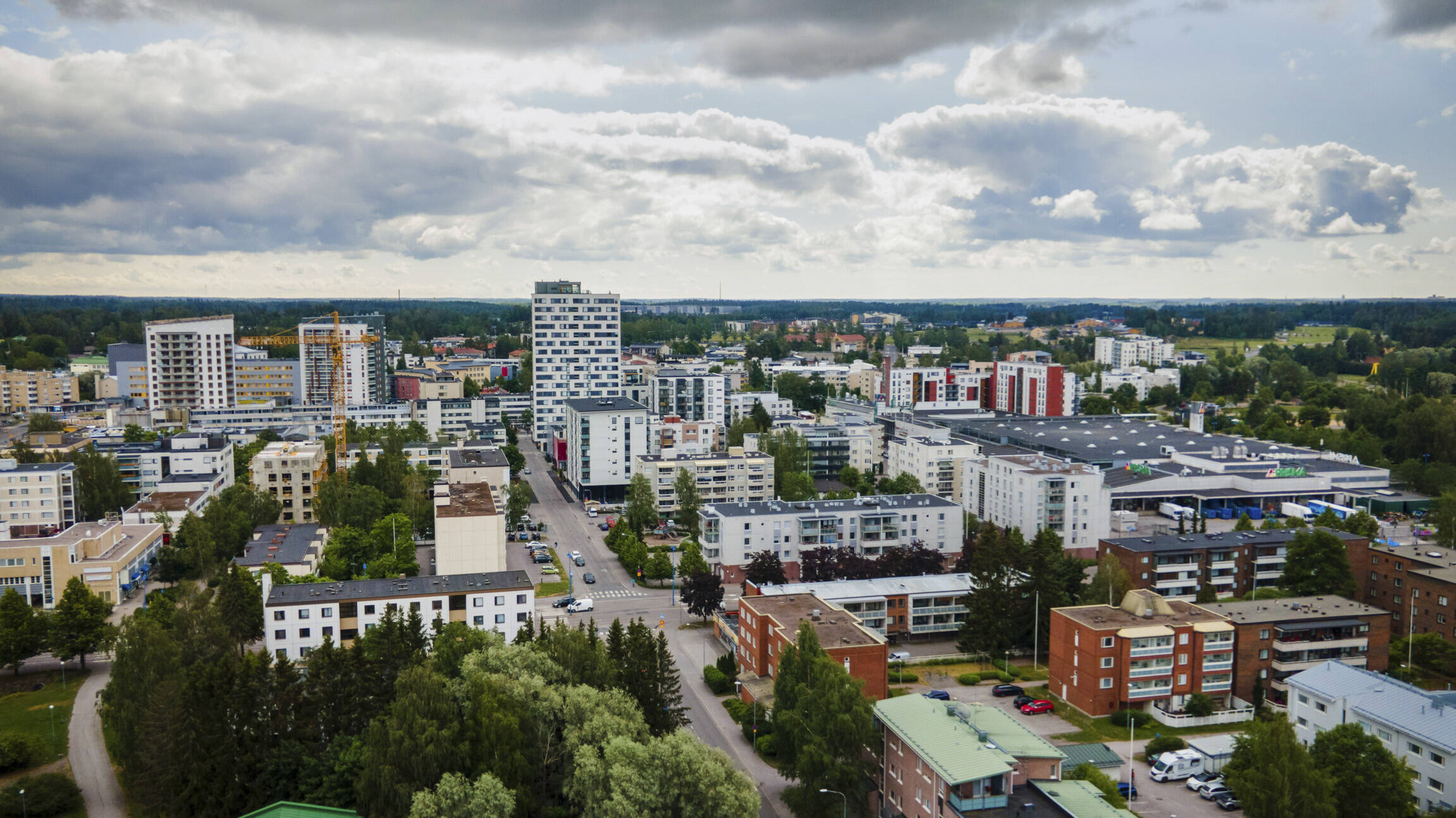 Ilmakuva Järvenpään keskustasta pilvisenä kesäpäivänä. Kuvassa näkyy tiivistä kaupunkirakennetta, kerrostaloja, liikekeskuksia ja vihreitä puistoalueita. Keskellä kohoaa useita korkeita asuinrakennuksia, ja etualalla näkyy matalampia punatiilisiä kerrostaloja puiden ympäröiminä. Taivaalla on suuria tummia pilviä, joiden välistä siivilöityy auringonvaloa kaupungin ylle.