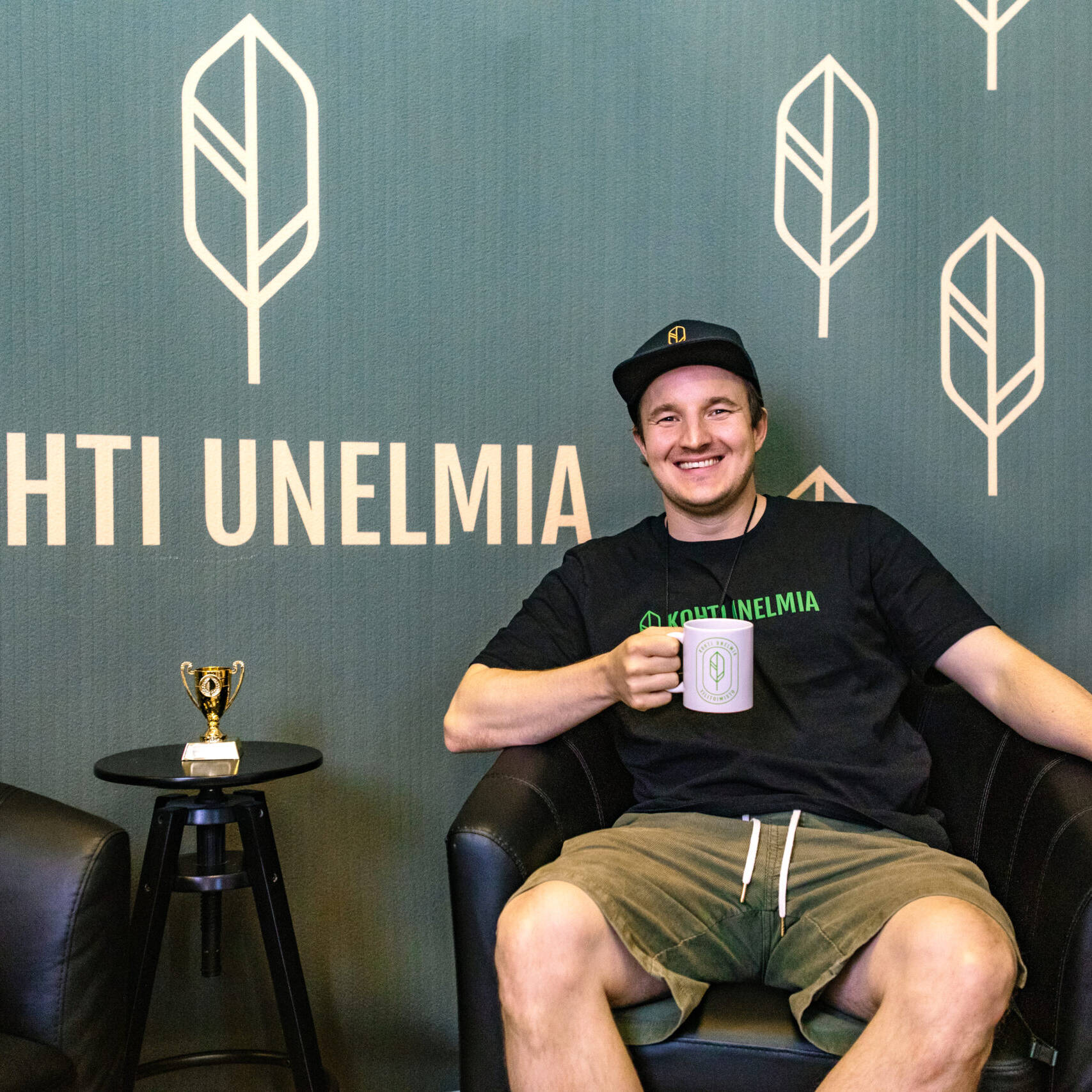 Johannes Laine istuu nojatuolissa Kohti Unelmia -logolla varustetun seinän edessä ja pitää kädessään yrityksen mukia.