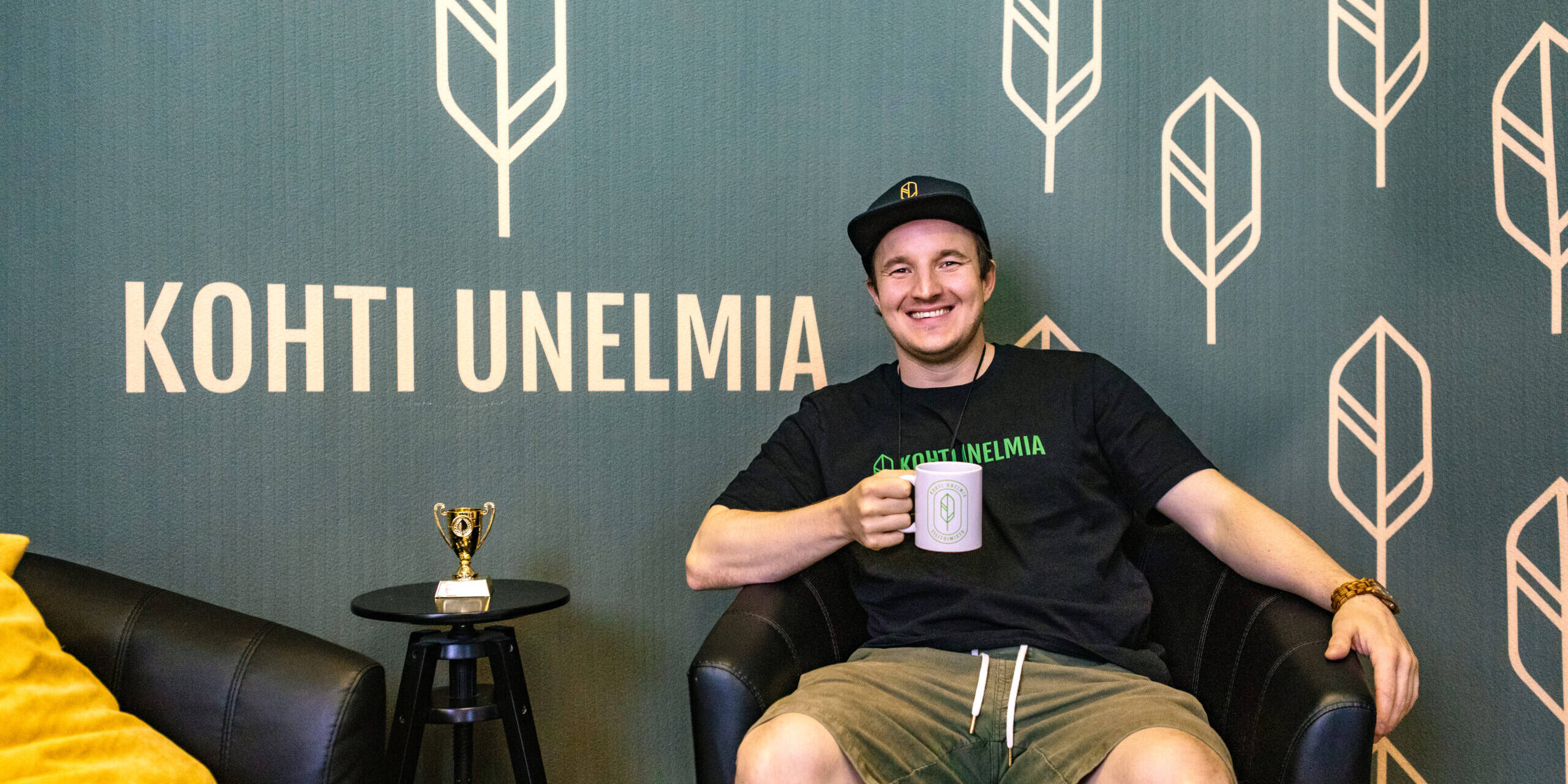 Johannes Laine istuu nojatuolissa Kohti Unelmia -logolla varustetun seinän edessä ja pitää kädessään yrityksen mukia.