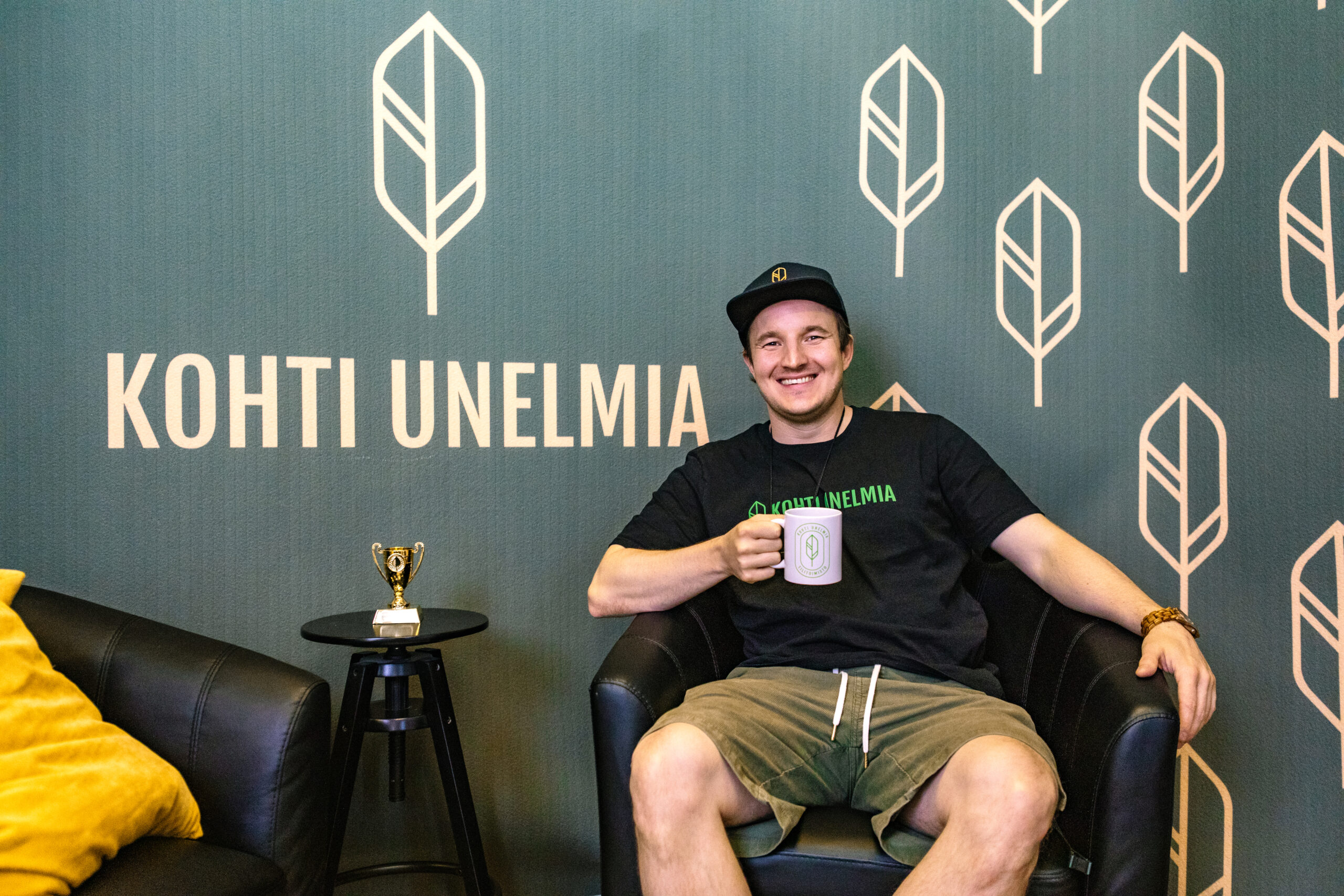 Johannes Laine istuu nojatuolissa Kohti Unelmia -logolla varustetun seinän edessä ja pitää kädessään yrityksen mukia.