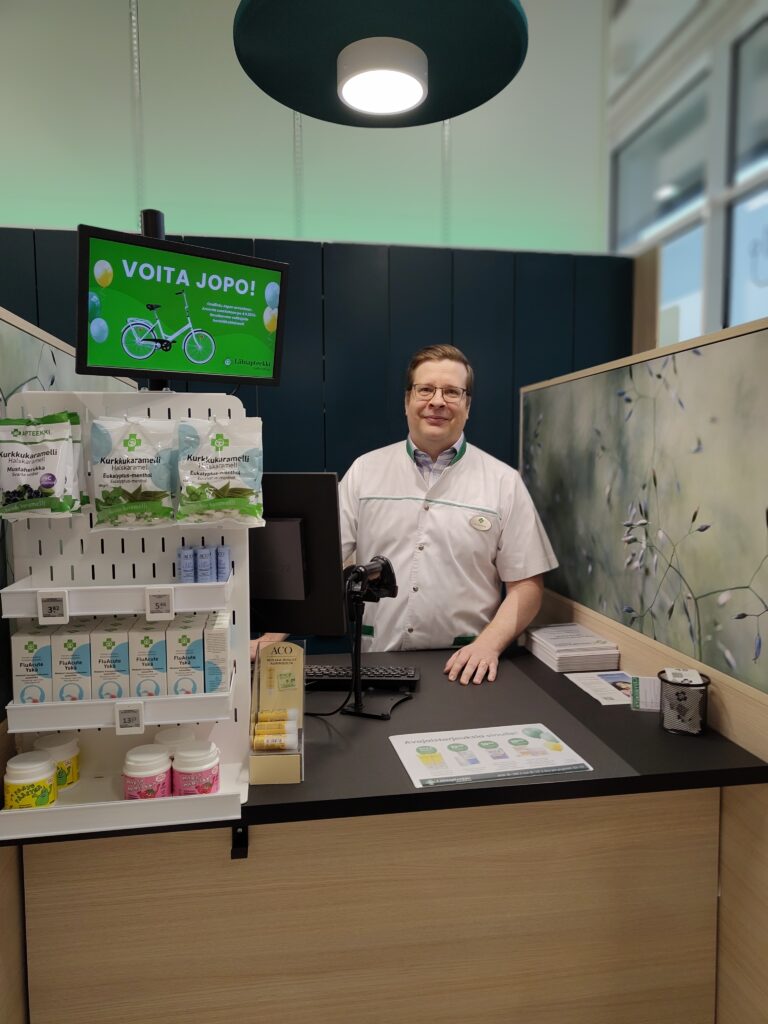 Apteekin työntekijä seisoo palvelutiskin takana Järvenpään Lähiapteekissa. Tiskin vieressä on apteekkituotteita ja taustalla näyttö, jossa mainostetaan polkupyörän arvontaa.