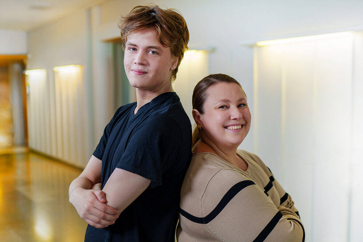 Nuori mies ja nainen seisovat selät vastakkain valoisassa käytävässä ja hymyilevät kameralle. Miehellä on musta t-paita ja naisella beige mustakuvioinen paita.