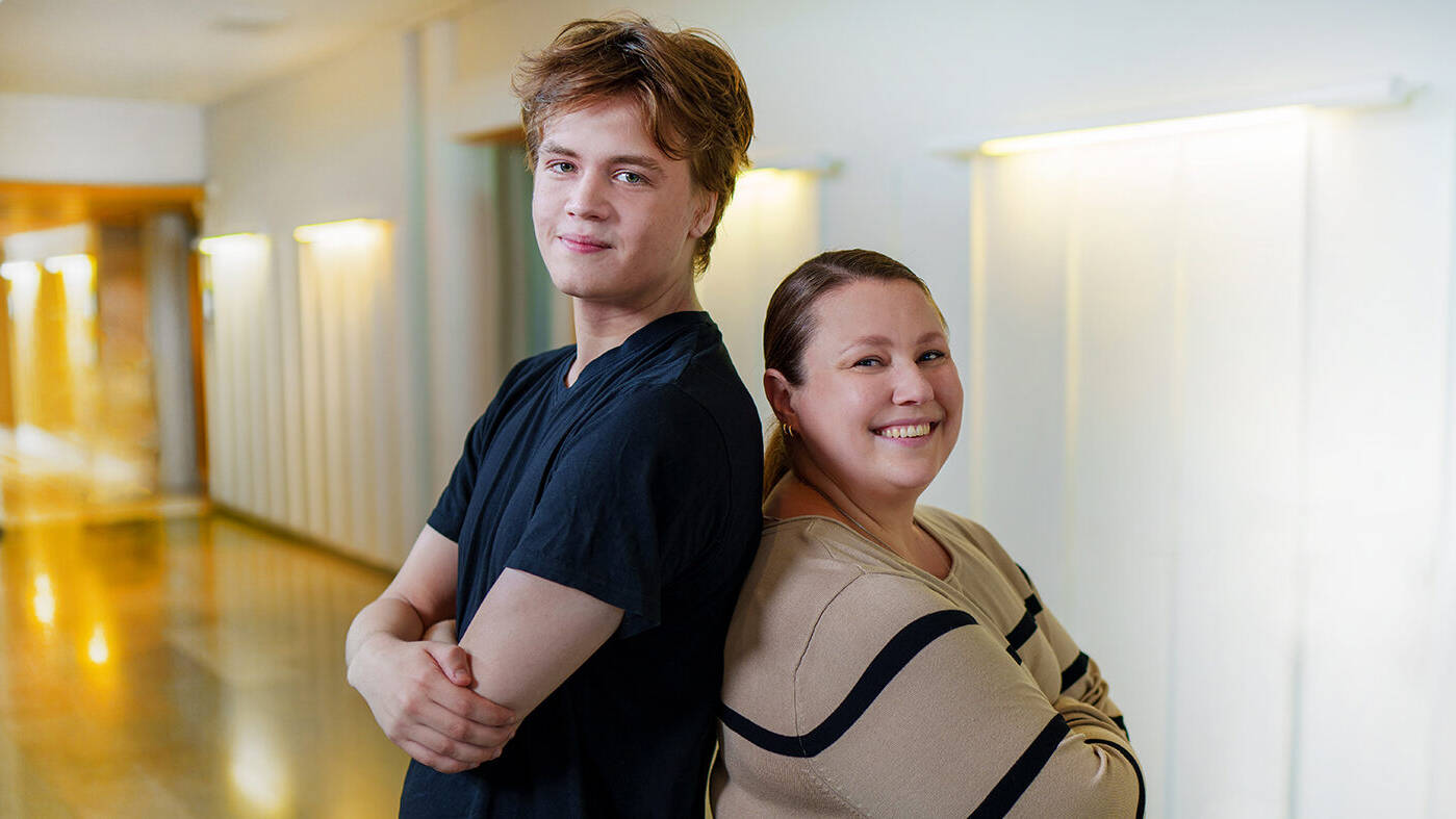 Nuori mies ja nainen seisovat selät vastakkain valoisassa käytävässä ja hymyilevät kameralle. Miehellä on musta t-paita ja naisella beige mustakuvioinen paita.