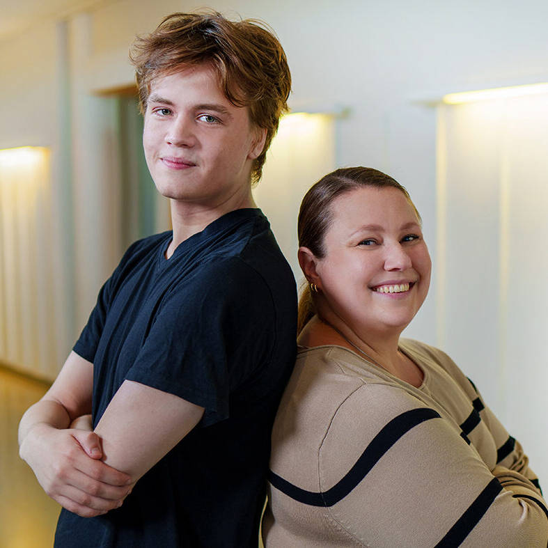 Nuori mies ja nainen seisovat selät vastakkain valoisassa käytävässä ja hymyilevät kameralle. Miehellä on musta t-paita ja naisella beige mustakuvioinen paita.