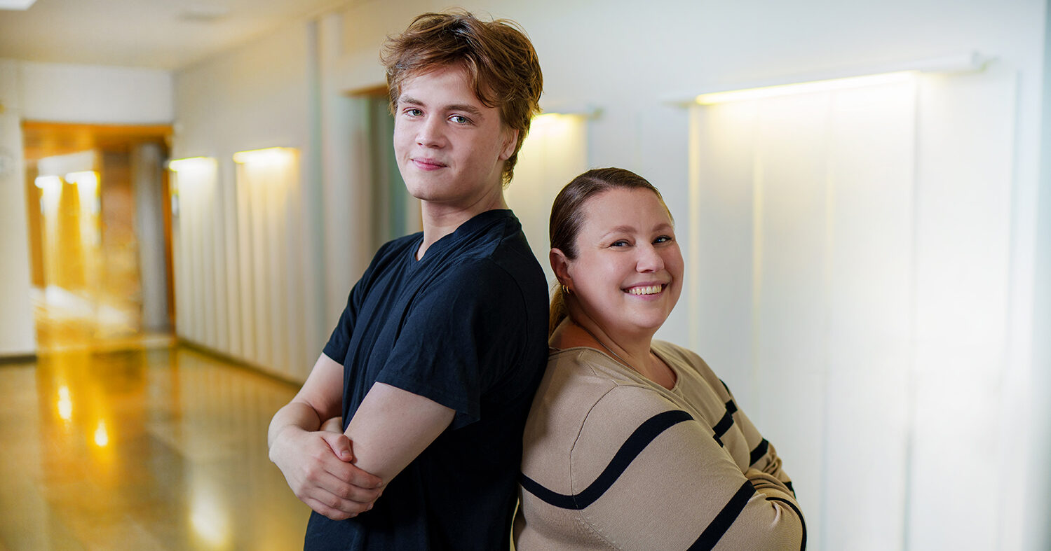 Nuori mies ja nainen seisovat selät vastakkain valoisassa käytävässä ja hymyilevät kameralle. Miehellä on musta t-paita ja naisella beige mustakuvioinen paita.