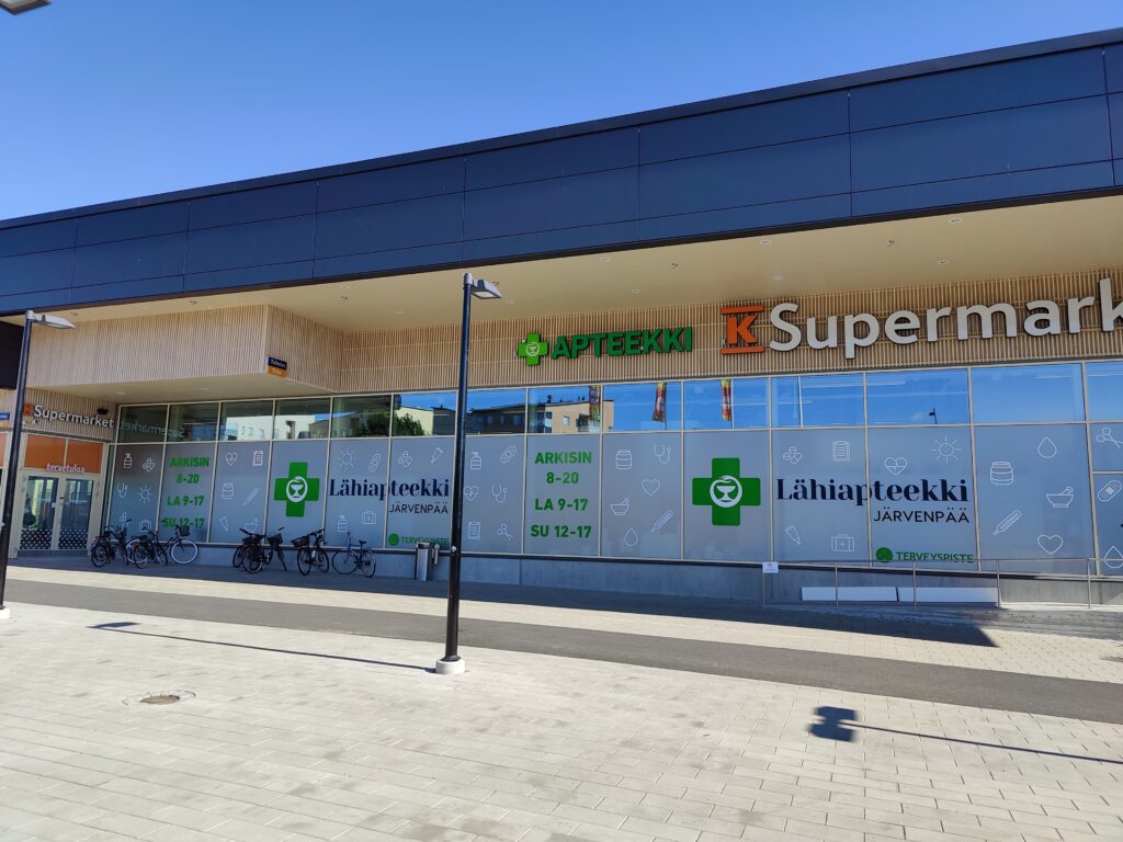 Järvenpään Lähiapteekin ja K-Supermarketin julkisivu Pajalan liikekeskuksessa aurinkoisena päivänä. Apteekin ikkunateippauksissa näkyvät Lähiapteekin tunnukset ja aukioloajat.