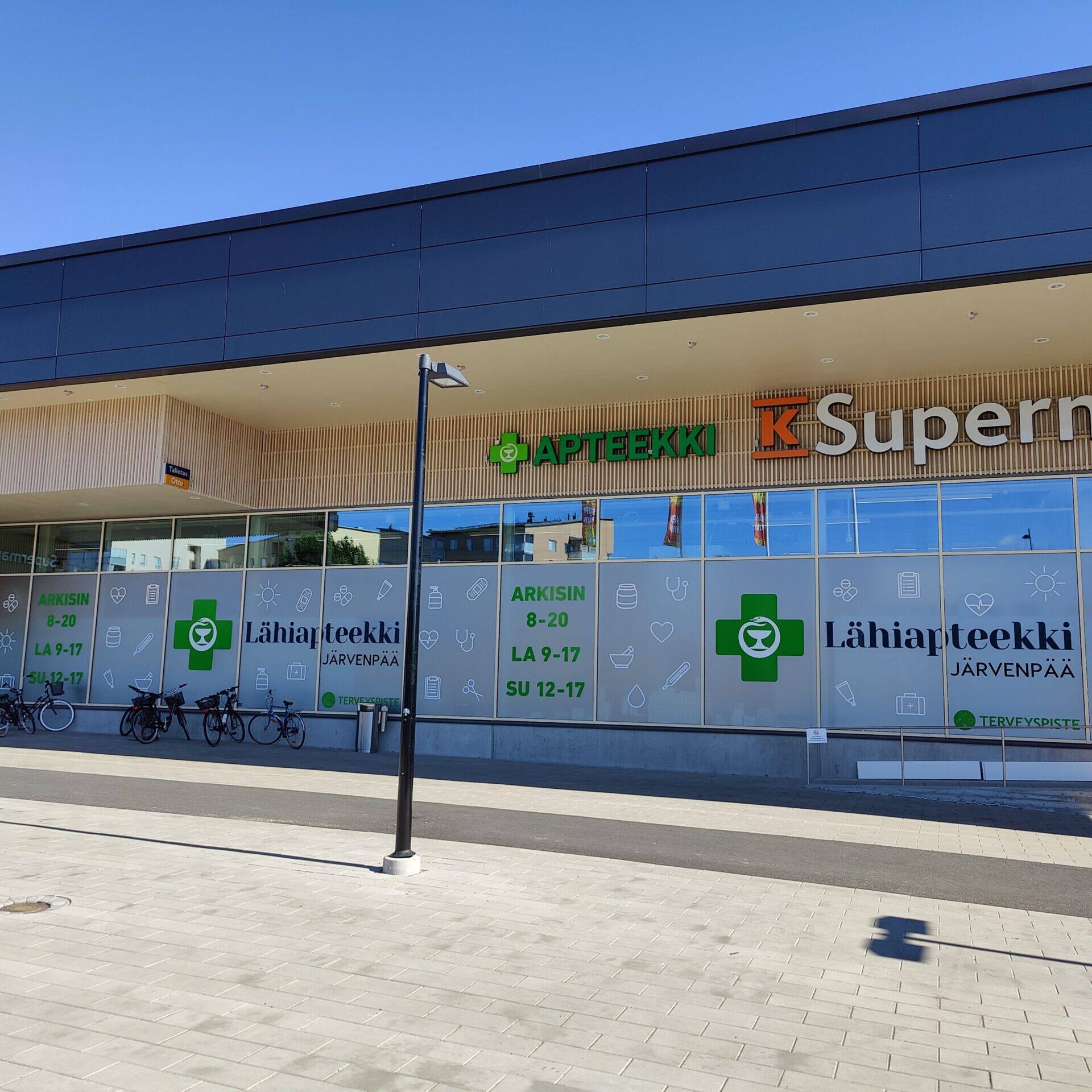 Järvenpään Lähiapteekin ja K-Supermarketin julkisivu Pajalan liikekeskuksessa aurinkoisena päivänä. Apteekin ikkunateippauksissa näkyvät Lähiapteekin tunnukset ja aukioloajat.