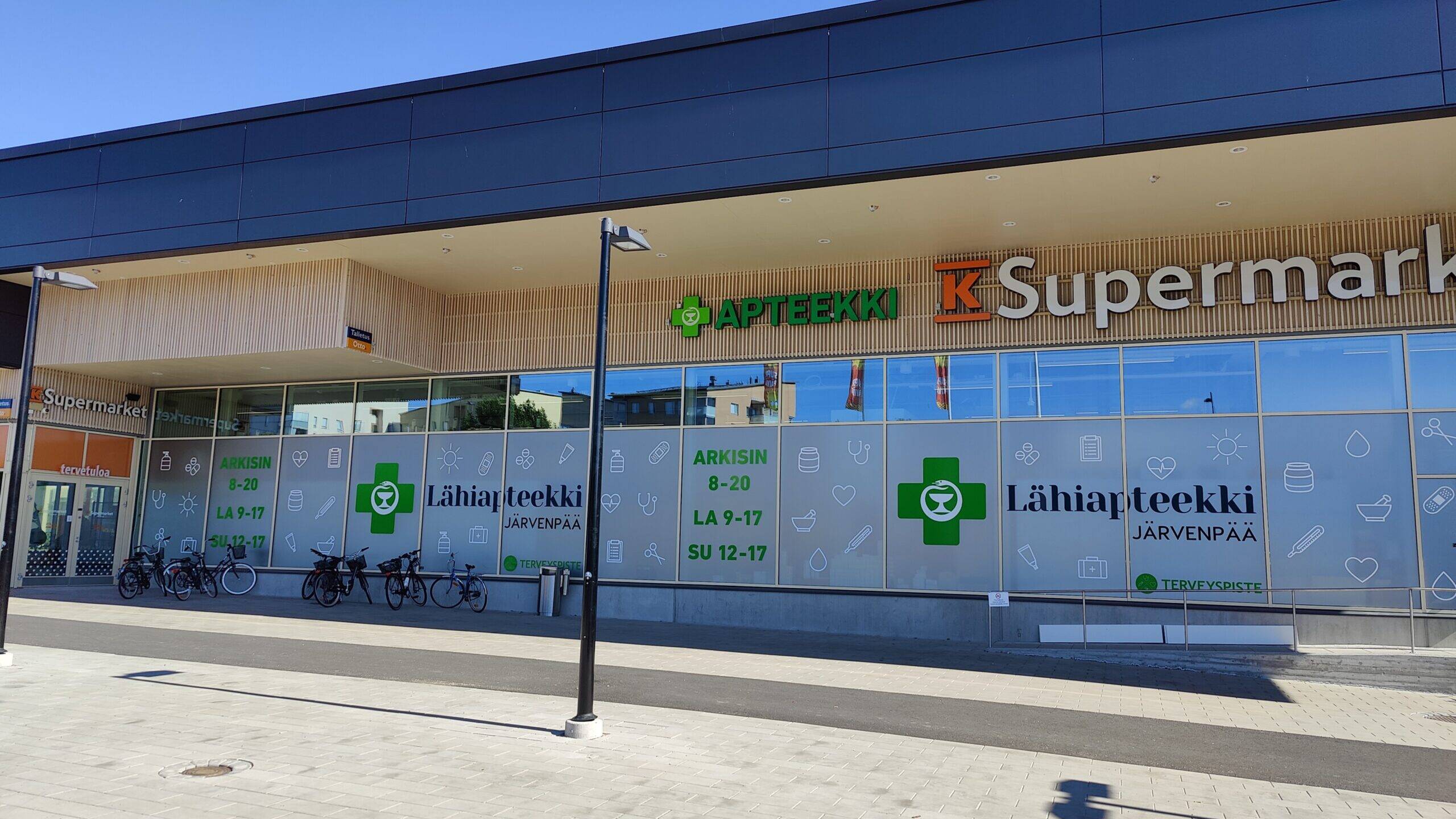 Järvenpään Lähiapteekin ja K-Supermarketin julkisivu Pajalan liikekeskuksessa aurinkoisena päivänä. Apteekin ikkunateippauksissa näkyvät Lähiapteekin tunnukset ja aukioloajat.