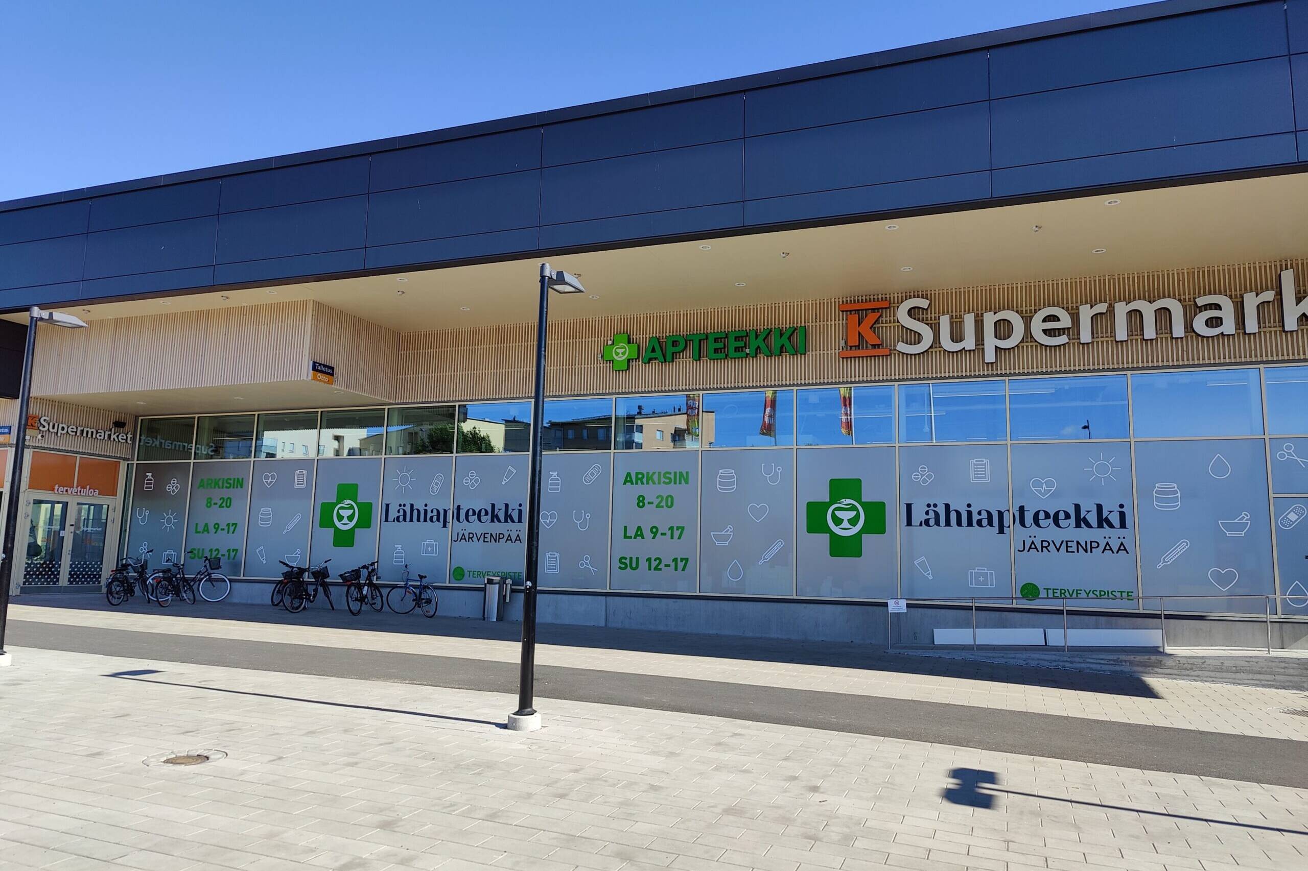 Järvenpään Lähiapteekin ja K-Supermarketin julkisivu Pajalan liikekeskuksessa aurinkoisena päivänä. Apteekin ikkunateippauksissa näkyvät Lähiapteekin tunnukset ja aukioloajat.